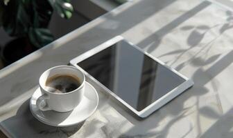 Tablet on a light blue table with a cup of tea on the right, shadow from window, top view photo
