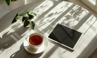 Tablet on a light blue table with a cup of tea on the right, shadow from window, top view photo