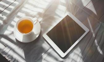 Tablet on a light blue table with a cup of tea on the right, shadow from window, top view photo