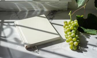 Closed notebook on a light gray table with a bunch of grapes on the right photo