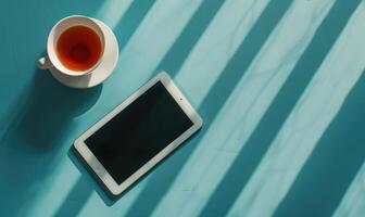 Tablet on a light blue table with a cup of tea on the right, shadow from window, top view photo
