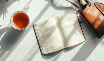 Closed notebook on a light gray table with a cup of tea on the left and a handbag on the right photo