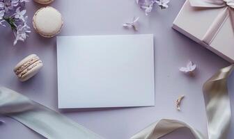 White blank card on a pastel lilac table, with a silk ribbon and a small box of macarons photo