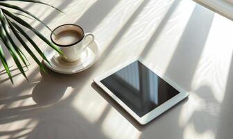 Tablet on a light blue table with a cup of tea on the right, shadow from window, top view photo