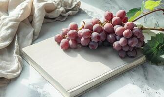 Closed notebook on a light gray table with a bunch of grapes on the right, soft window light photo