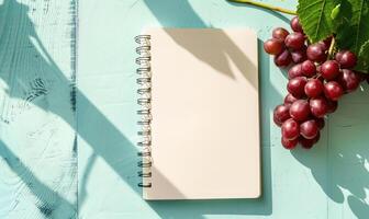 Closed notebook on a light blue table with a bunch of grapes on the left photo