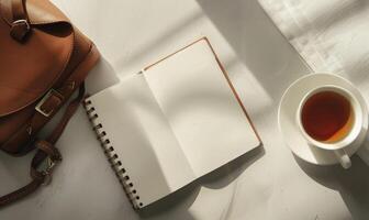 Closed notebook on a light gray table with a cup of tea on the right and a handbag on the left photo