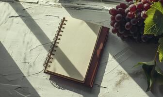 Closed notebook on a light gray table with a bunch of grapes on the right photo