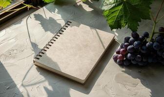 Closed notebook on a light gray table with a bunch of grapes on the right photo