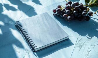 Closed notebook on a light blue table with a bunch of grapes on the right, shadow from window photo