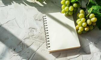 Closed notebook on a light gray table with a bunch of grapes on the right, soft window light photo