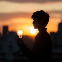 silhouette of young man holding light bulb in front of city at sunset photo