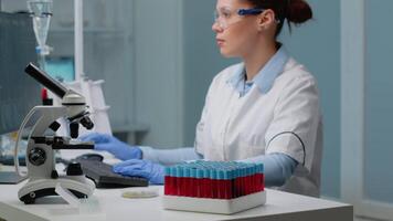 Microbiology specialist using computer in laboratory while having vacutainers tray with fluid blood samples and microscope prepared for genetic examination. Chemist woman in lab coat video