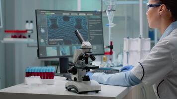 Biochemistry doctor analyzing vacutainer with blood while having microscope on desk and using computer in professional medical laboratory. Specialist woman doing analysis experiment video