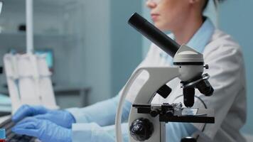 Scientific woman with microscope tool studying petri dish while using computer in background. Biology doctor in laboratory with research equipment and instruments for science innovation video