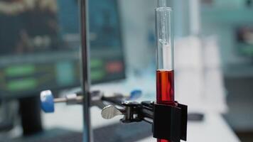Close up of test tube filled with blood or dna liquid connected to dripping pipe in laboratory. Chemical glassware and flask with red solution on desk used for development industry video
