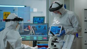 squadra di scienziati nel coveall discutere di virus sviluppo nel davanti di pc e tavoletta nel attrezzata laboratorio. medico cose analizzando vaccino Evoluzione utilizzando alto Tech per ricercando trattamento video