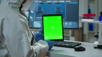Chemist man wearing protection suit holding tablet with green mockup in modern equipped lab. Team of microbiologists doing vaccine research writing on device with chroma key, isolated display. video