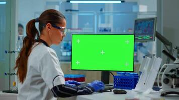 Woman researcher typing at computer with green screen mockup display, template in applied science laboratory. Engineer conducting experiments in background, examining vaccine evolution using high tech video