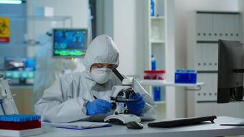 Medical research scientist conducting DNA experiments under microscope wearing protection suit and writing informations on clipboard. Chemist using high tech for vaccine development against covid19 video