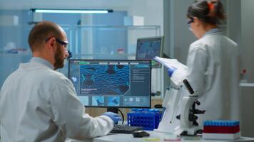 Man scientist working to develop a new vaccine in modern equipped laboratory, typing at computer. Group of biochemistry doctors examining virus evolution using high tech for researching treatment video