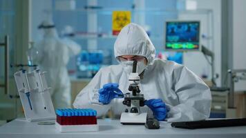 Scientist in ppe suit making adjustments and looking through laboratory microscope, typing on pc. Chemist in coverall working with various bacteria, tissue blood samples for antibiotics research video