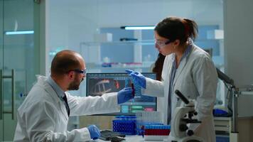 Laboratory medical stuff disscussing about vaccine development holding test tube with blood sample. Medical scientist working with DNA scan image in modern equipped laboratory using high tech video