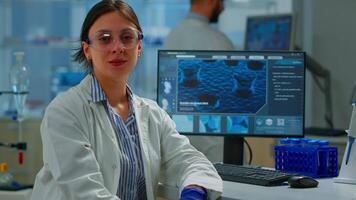 Chemist with lab coat sitting in laboratory looking at camera smiling, while colleague working on computer in background. Scientist examining virus evolution using high tech for scientific research video