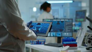 Close up of scientist in lab coat working holding tablet standing in modern equipped laboratory. Team chemists examining vaccine evolution using high tech and chemistry tools for virus development video