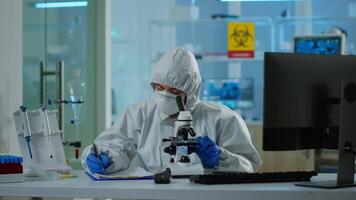 Biologist in ppe suit working on microscope and writing information on clipboard in equipped laboratory. Chemist examining vaccine evolution using high tech researching diagnosis against covid19 virus video
