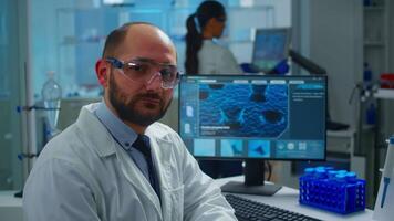 Portrait of exhausted scientist man looking at camera sitting in front of computer with DNA scan image. Chemist examining virus evolution using high tech and chemistry tools for scientific research video