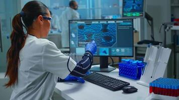 mulher tecnólogo fazendo uma laboratório teste examinando uma frasco com sangue amostra, químico segurando tubo com líquidos dentro. cientista trabalhando com vários bactérias lenço de papel e dna Varredura imagem em computador video