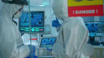 Chemists in coverall trying to develop vaccine using tablet standing behind the glass wall working in danger area of lab. Scientists examining virus evolution researching for treatment against covid19 video