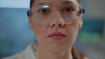 Biochemist with safety goggles and white coat in laboratory, face of scientist looking at camera while she works on chemistry development in lab. Portrait of woman doing research. Close up video