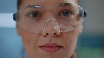 Biochemical engineer wearing protective glasses in science lab. Chemistry specialist with goggles smiling at camera while she works on research development in laboratory. Close up video