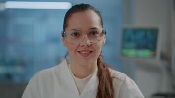 Portrait of woman scientist with goggles smiling in laboratory, getting ready to work on science experiment and development. Microbiology researcher wearing safety glasses in lab. Close up video