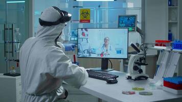 Lab technician in ppe suit sitting at table in medical research laboratory, holding test tube and talking on call with senior doctor giving online advices during virtual meeting. video