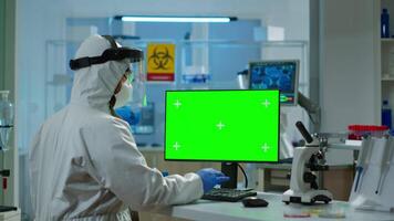 Scientist man wearing protection suit typing on computer with green mockup in modern equipped lab. Team of microbiologists doing vaccine research writing on device with chroma key, isolated display. video