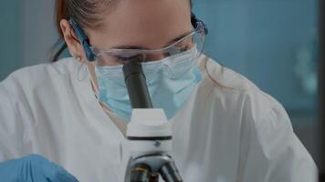 Biologist with face mask using microscope to work on experiment in science laboratory. Woman working with microscopic lens and magnifying glass on scientific tool, wearing lab protection video