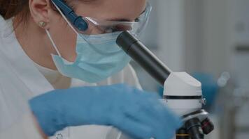 Specialist analyzing dna on microscope in laboratory, wearing face mask, glasses and gloves for protection. Science engineer using magnifying glass on optical microscopic tool. Close up video