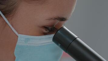 Biologist with face mask using microscopic lens on chemistry tool in laboratory. Science engineer using microscope with magnifying glass and selective focus to work on lab experiment. video