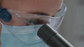 chimico guardare attraverso microscopico lente su ottico attrezzo, indossare viso maschera e protettivo occhiali nel laboratorio. microbiologo utilizzando microscopio e selettivo messa a fuoco per sperimentare. vicino su video