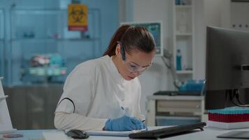 Woman chemist taking notes for science research in laboratory, working on development. Scientist using computer in lab to work on experiment and write on paper for biology expertise. video