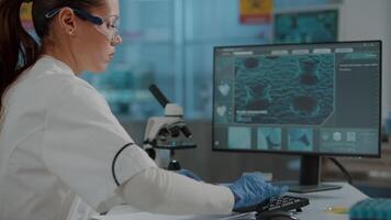 Microbiologist taking notes on paper and analyzing dna on computer, wearing protective glasses and gloves in laboratory. Scientist using monitor to develop chemistry experiment in lab. video