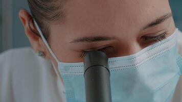 Woman scientist using microscope with magnifying glass for science experiment in laboratory. Specialist wearing face mask, working with microscopic lens on scientific tool. Close up video