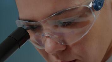 Woman chemist with protective glasses using microscope in laboratory. Microbiologist working with microscopic lens on optical tool to analyze genetic substance with magnifying glass. Close up video