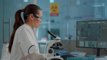 Science engineer using microscope to analyze dna sample in laboratory. Woman biologist working with microscopic glass lens on lab tool and computer, wearing protective goggles and gloves. video