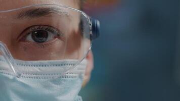 Scientist showing one eye on camera in laboratory, wearing safety glasses and face mask. Woman biologist looking at camera with face of face, working in research lab. Close up video