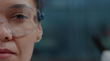 Woman scientist wearing protective glasses with virtual reality in science laboratory. Chemistry specialist with futuristic goggles for simluation, showing one eye on camera. Close up video