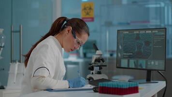 Biology researcher using computer and taking notes for science experiment in laboratory. Woman chemist working on biotechnology expertise with lab equipment, writing diagnosis on paper. video